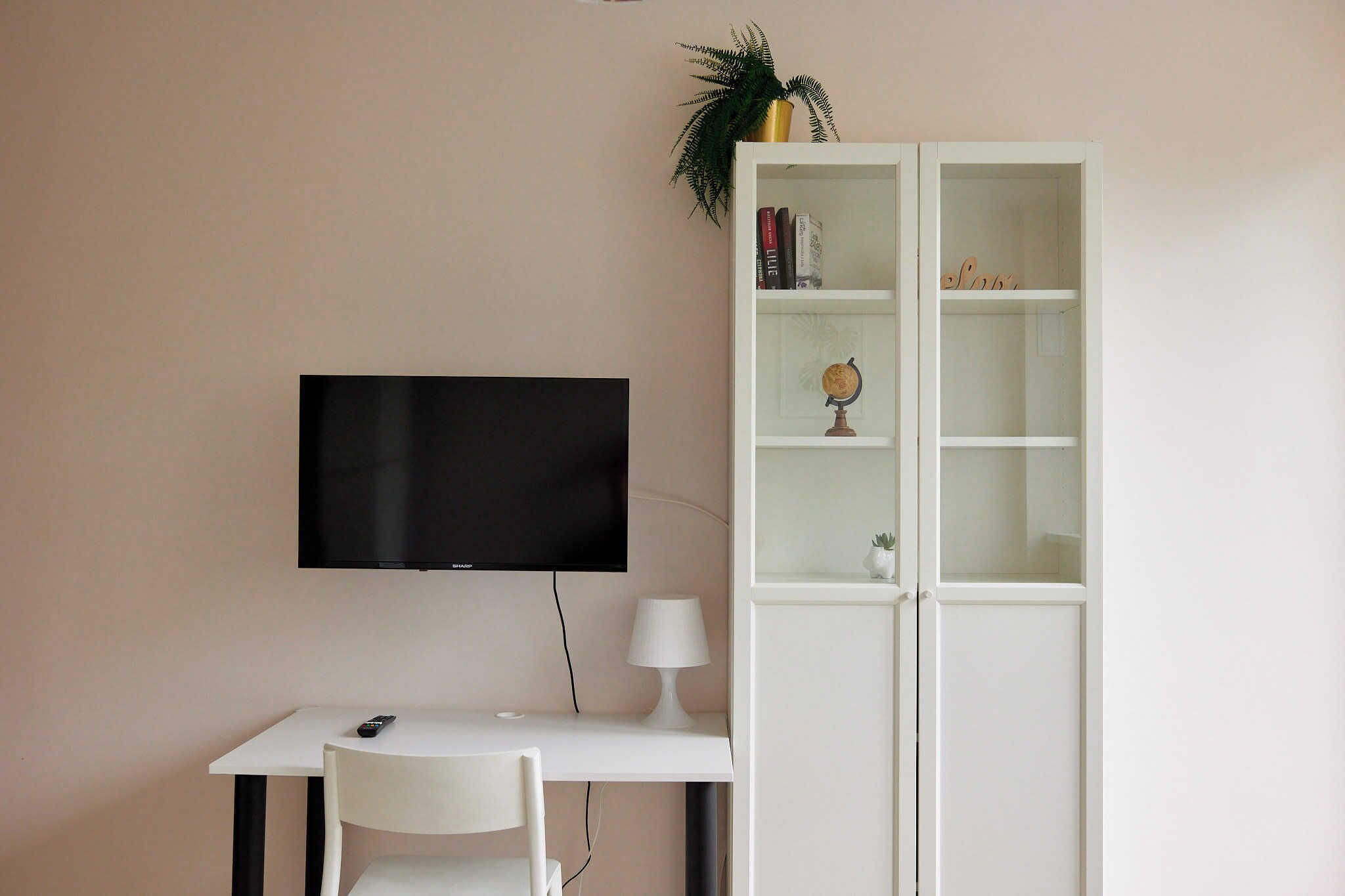 A room with a flat-screen TV mounted on the wall, a white desk with a chair, a white lamp, and a white cabinet with shelves.