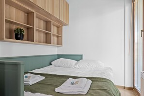 Ein Schlafzimmer mit einem Bett, einer grünen Decke, weißen Kissen, einem hölzernen Regal mit einem Kakteen und einem Fenster mit Jalousien.