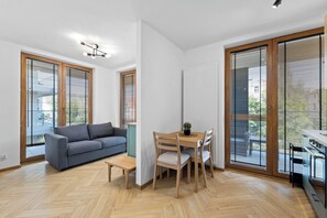 Un salon moderne avec un canapé gris, une table à manger en bois et des chaises. De grandes fenêtres sont équipées de stores, et on y trouve un luminaire au plafond ainsi qu’un radiateur.