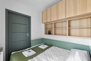 Quarto com uma cama, uma porta cinzenta e uma prateleira de madeira acima da cama.