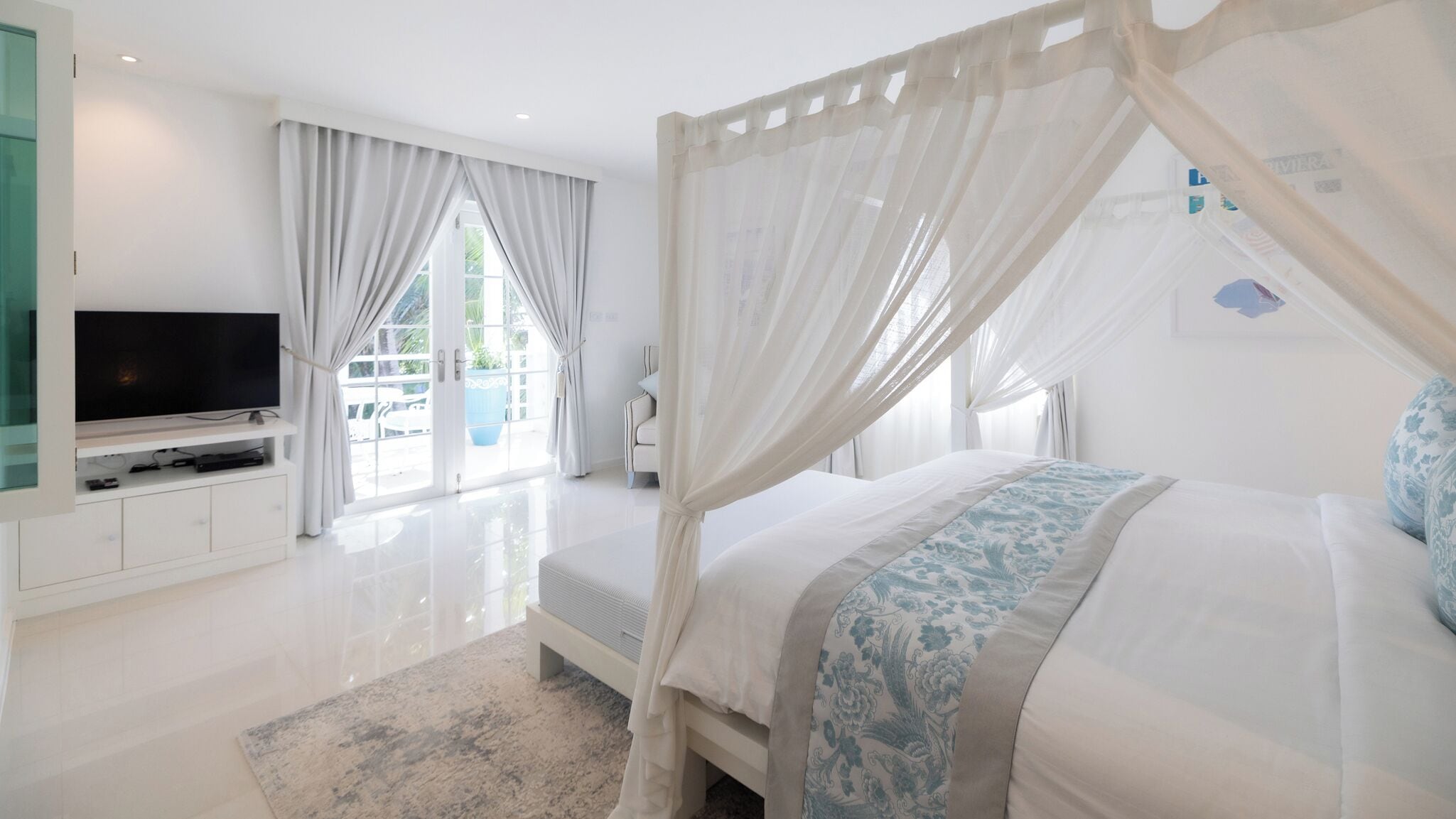Un dormitorio con una cama con dosel, un televisor sobre un soporte y un ventanal con cortinas.