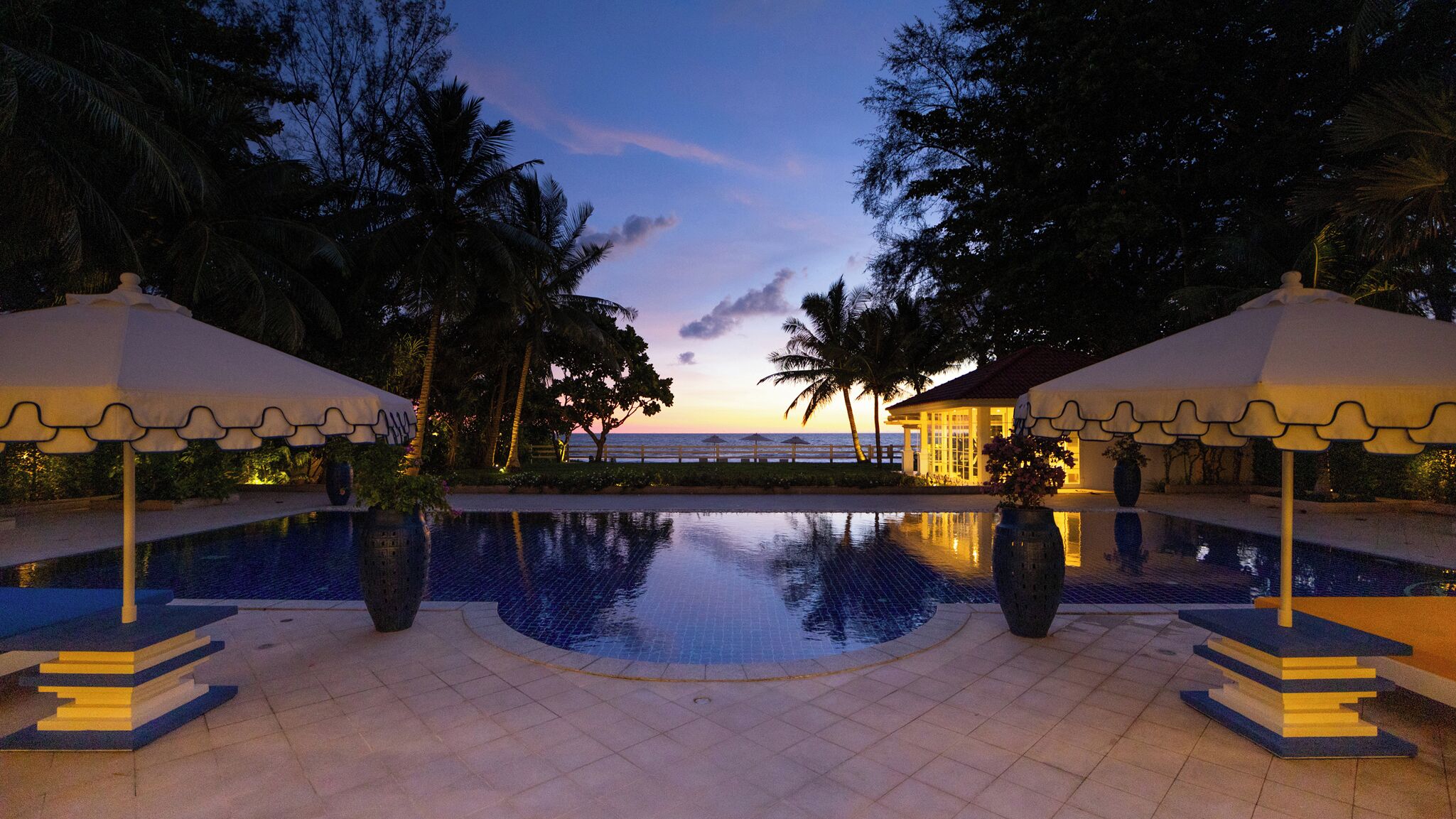 A swimming pool with a view of the sea at dusk, surrounded by palm trees and umbrellas.