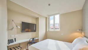 Et hotelværelse med en seng, et skrivebord med stol, telefon, fjernsyn og et vindue med udsigt til bygninger.