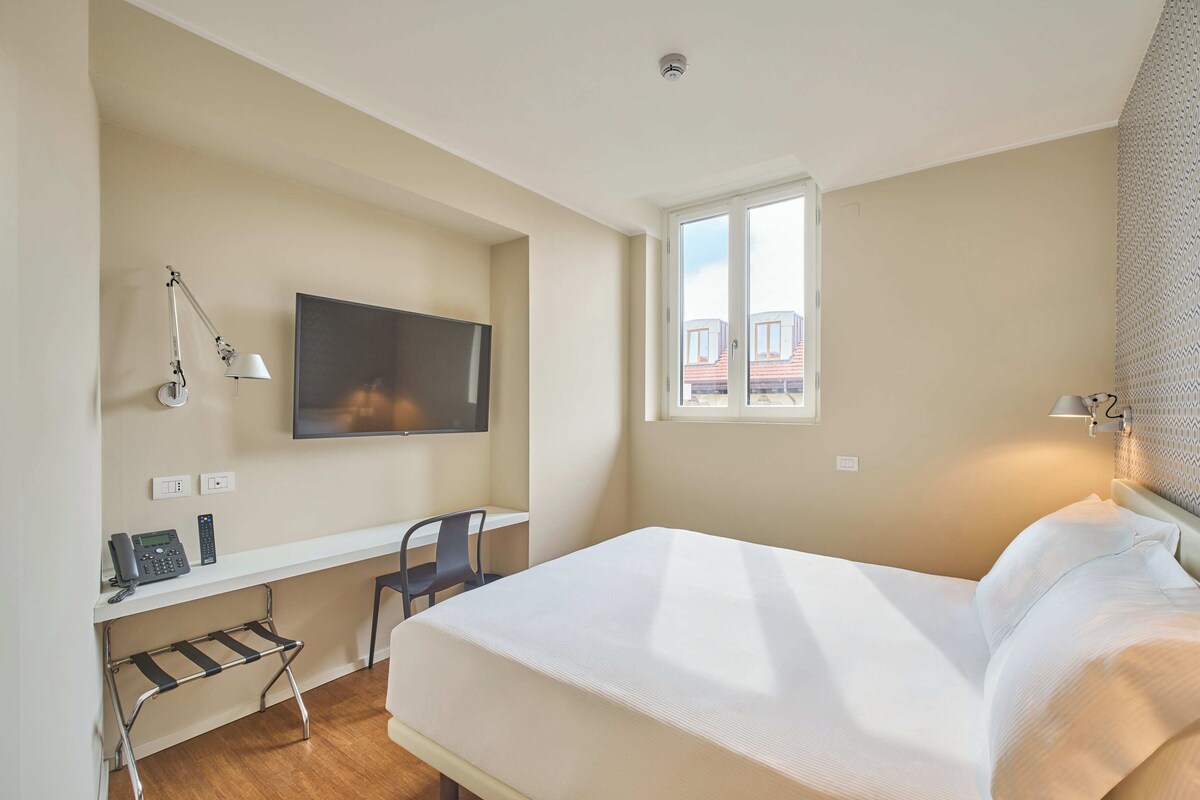 Ein Hotelzimmer mit einem Bett, einem Schreibtisch mit Stuhl, einem Telefon, einem Fernseher und einem Fenster mit Blick auf Gebäude.