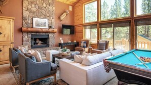 Una sala de estar acogedora con chimenea de piedra, asientos cómodos, mesa de billar y amplios ventanales que ofrecen vistas al bosque circundante.