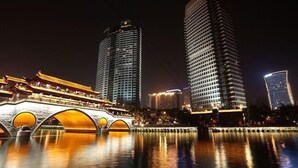 Une vue nocturne d’une ville, avec des bâtiments illuminés et un pont traditionnel enjambant une étendue d’eau.