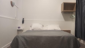 A single bed with a grey bedspread, a wooden nightstand with a remote control, and a black curtain. - Hotel Residence Saint Ouen (Saint-Ouen)