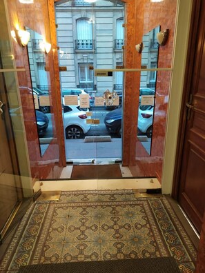 An entrance with a patterned floor, a glass door reflecting a street view, and a building with a sign 'poussez'. - Hotel Residence Saint Ouen (Saint-Ouen)