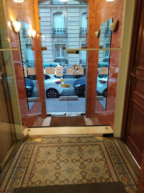An entrance with a patterned floor, a glass door reflecting a street view, and a building with a sign 'poussez'. - Hotel Residence Saint Ouen (Saint-Ouen)