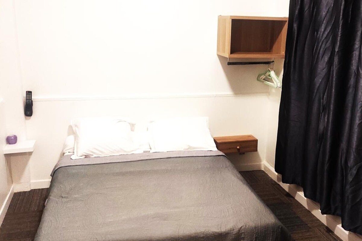 A small, single-bed room with a wooden nightstand, a bedside table, a hanging lamp, and a black curtain.