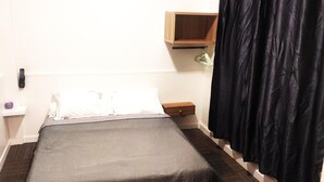 A small, single-bed room with a wooden nightstand, a bedside table, a hanging lamp, and a black curtain. - Hotel Residence Saint Ouen (Saint-Ouen)