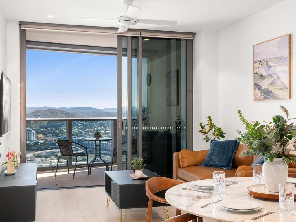 Een moderne woonkamer met een grote schuifpui naar een balkon, een eethoek met een tafel voor twee personen en een bank met decoratieve planten.