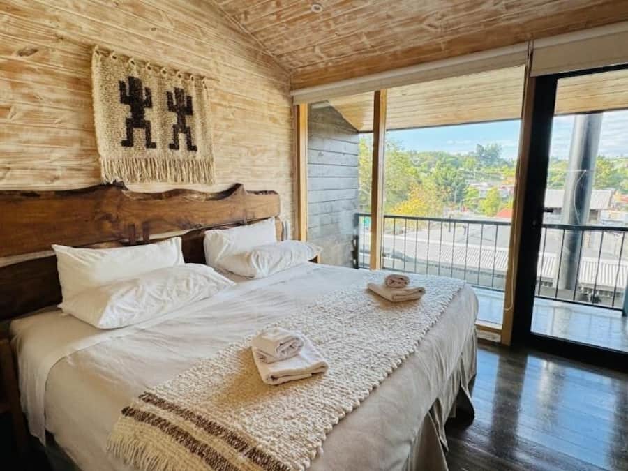 Un dormitorio con cabecera de madera, una cama grande con ropa blanca, un plaid tejido y vistas al exterior a través de un ventanal corredero.