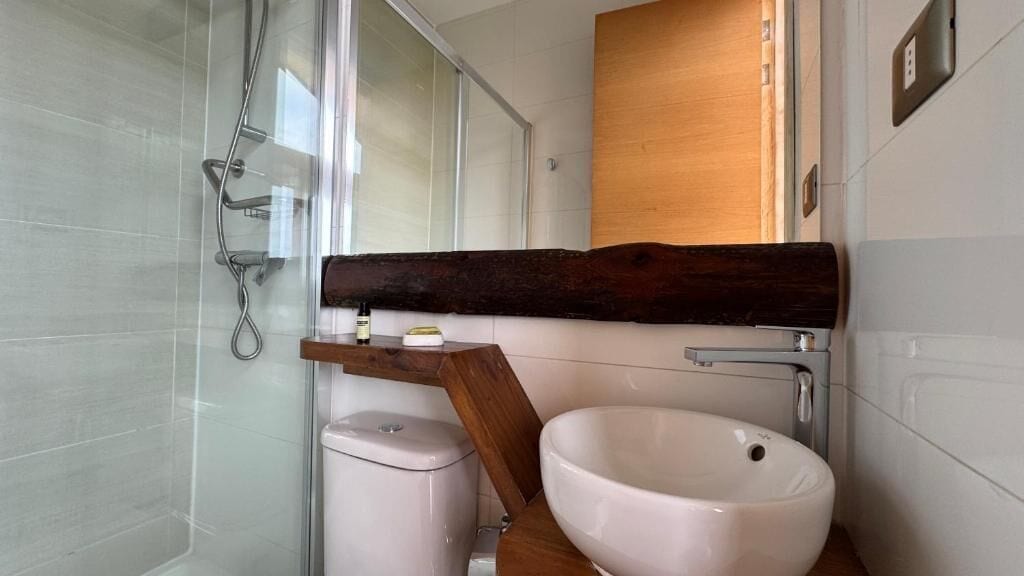 Un baño con cabina de ducha de vidrio, inodoro blanco, tocador de madera con lavabo y espejo.