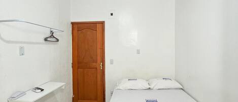 Una habitación pequeña y limpia con una cama, una puerta de madera y un estante blanco.