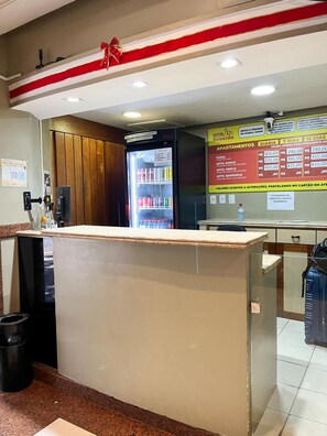 A hotel reception area with a counter, a refrigerator stocked with beverages, and a menu board displaying room rates. - Hotel Avenida (Belém)
