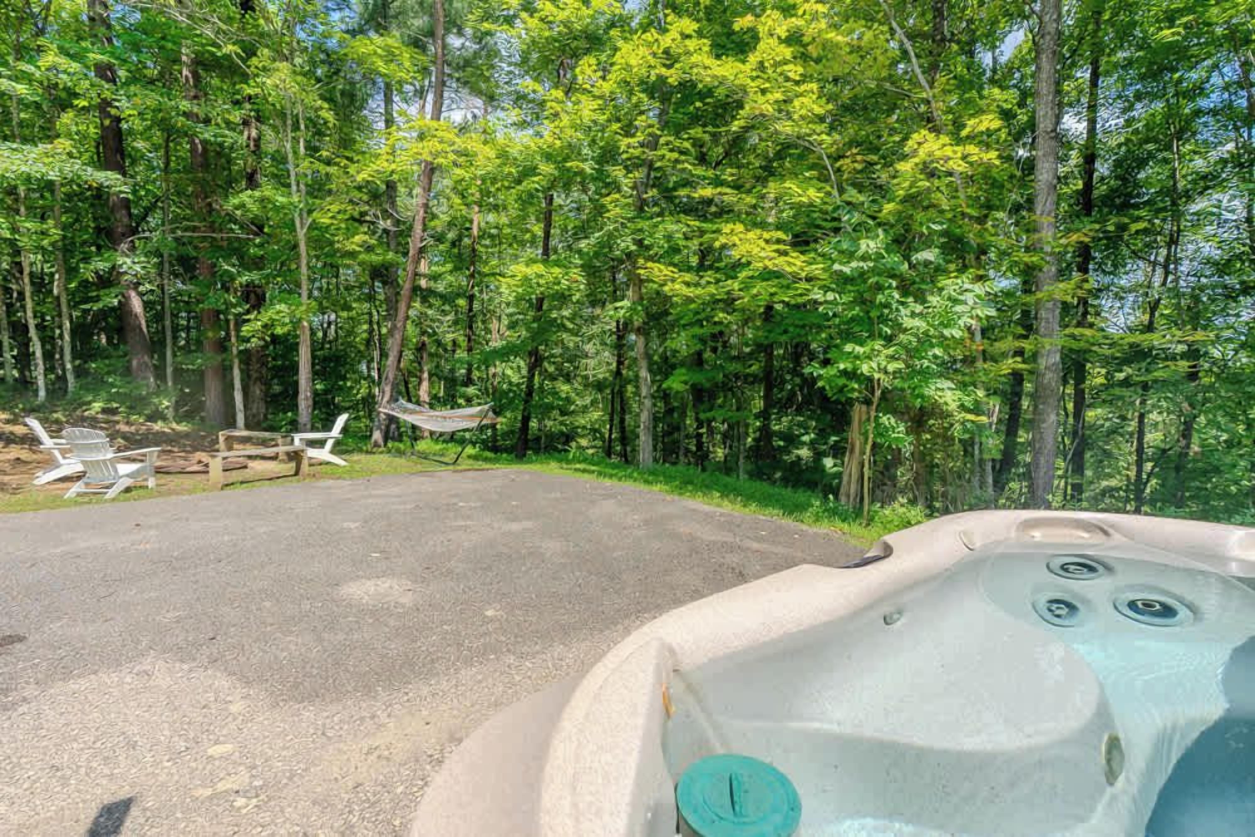 A hot tub in a forested area with a paved path and outdoor seating.