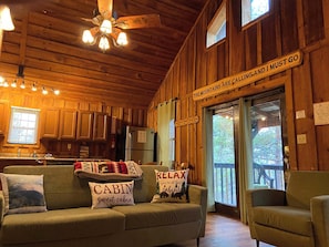 A cabin interior with a wooden ceiling, a kitchenette, a sofa with cabin-themed pillows, and a sign on the wall. - Pine Cove - Hot Tub - Pet Friendly - Large Yard (Campton)