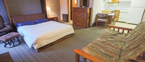 Une chambre à coucher avec un lit, une commode en bois, une chaise, une petite table et, au fond, un coin cuisine.