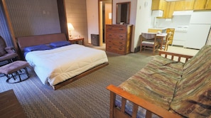 Un dormitorio con cama, cómoda de madera, silla, una mesita y una kitchenette al fondo.