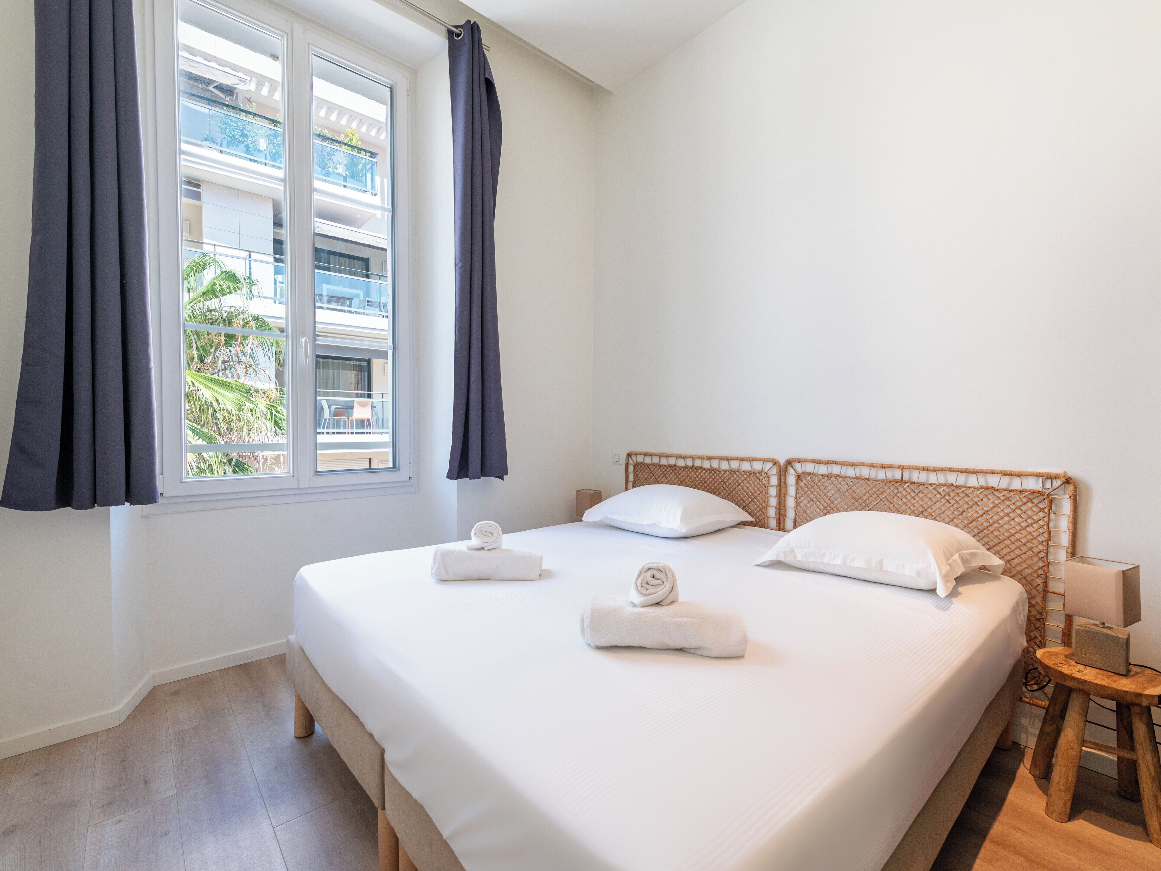 Ein Hotelzimmer mit einem Bett, einem Fenster mit Vorhängen, einem kleinen Holzschemel und einem Nachttisch.