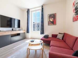 Een moderne woonkamer met een rode bank, een klein rond tafeltje, een televisie en kunstwerken aan de muur.
