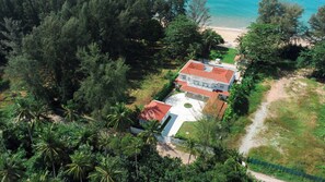 Ein großes, mehrstöckiges Haus mit rotem Dach, umgeben von dichtem tropischen Grün und einem Strand im Hintergrund.