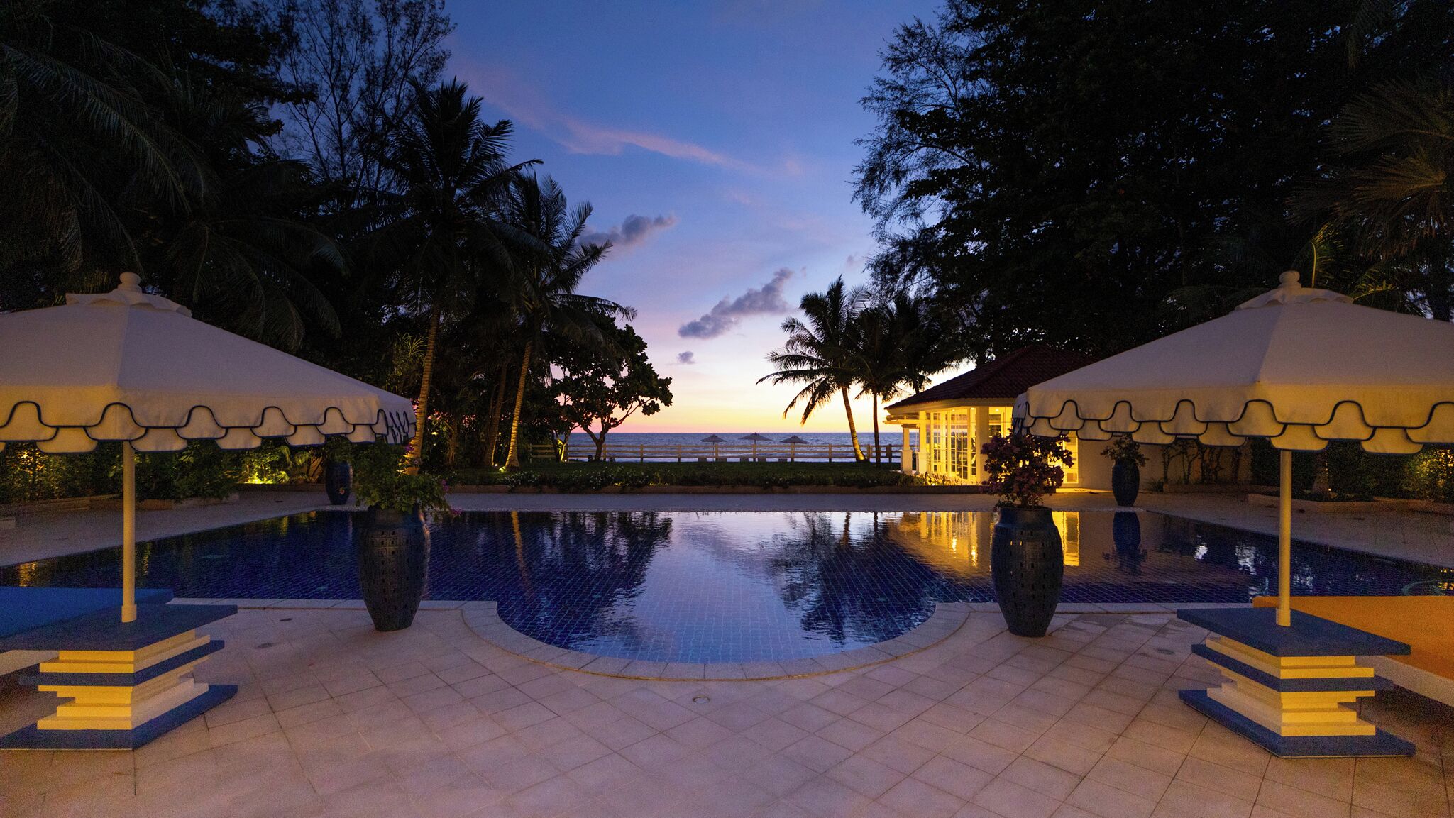 A pool area with a view of the sunset, palm trees, and a building with a white roof.