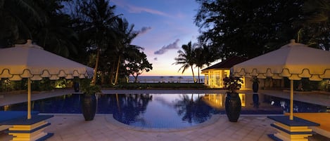 A pool area with a view of the sunset, palm trees, and a building with a white roof.
