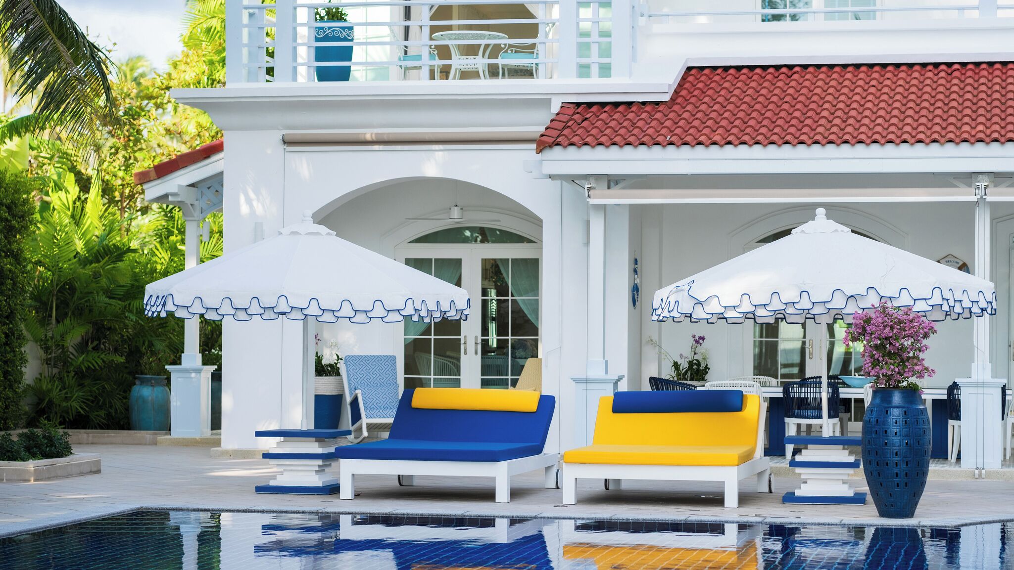 A poolside area with lounge chairs and umbrellas, a covered seating area, and a view of a building with a red-tiled roof.