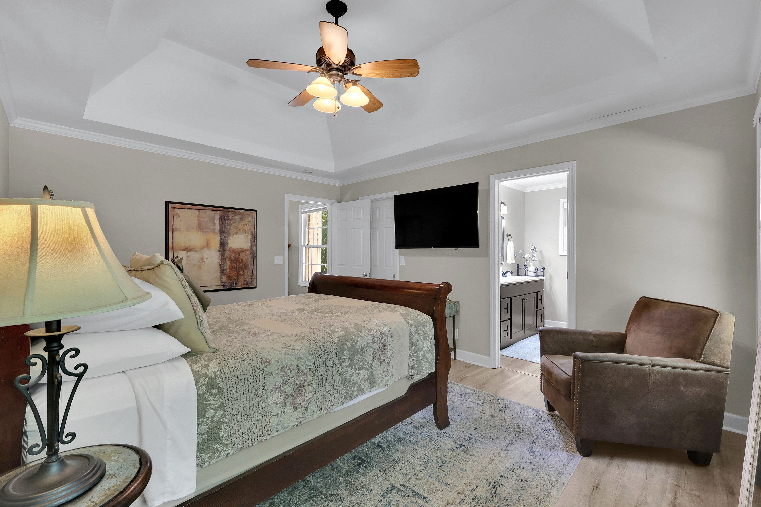 A bedroom with a bed, a chair, a ceiling fan, a TV, and a painting on the wall.