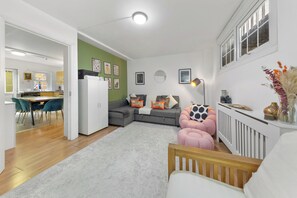 Un salon moderne avec un canapé d’angle gris, un pouf rose et une chaise en bois. On aperçoit une cuisine à l’arrière-plan, avec un coin repas.