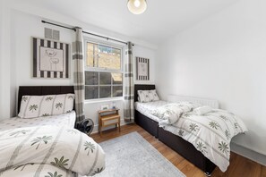 Ein Schlafzimmer mit zwei Betten, einem Fenster mit Vorhängen, einem Heizkörper und einem kleinen Nachttisch.