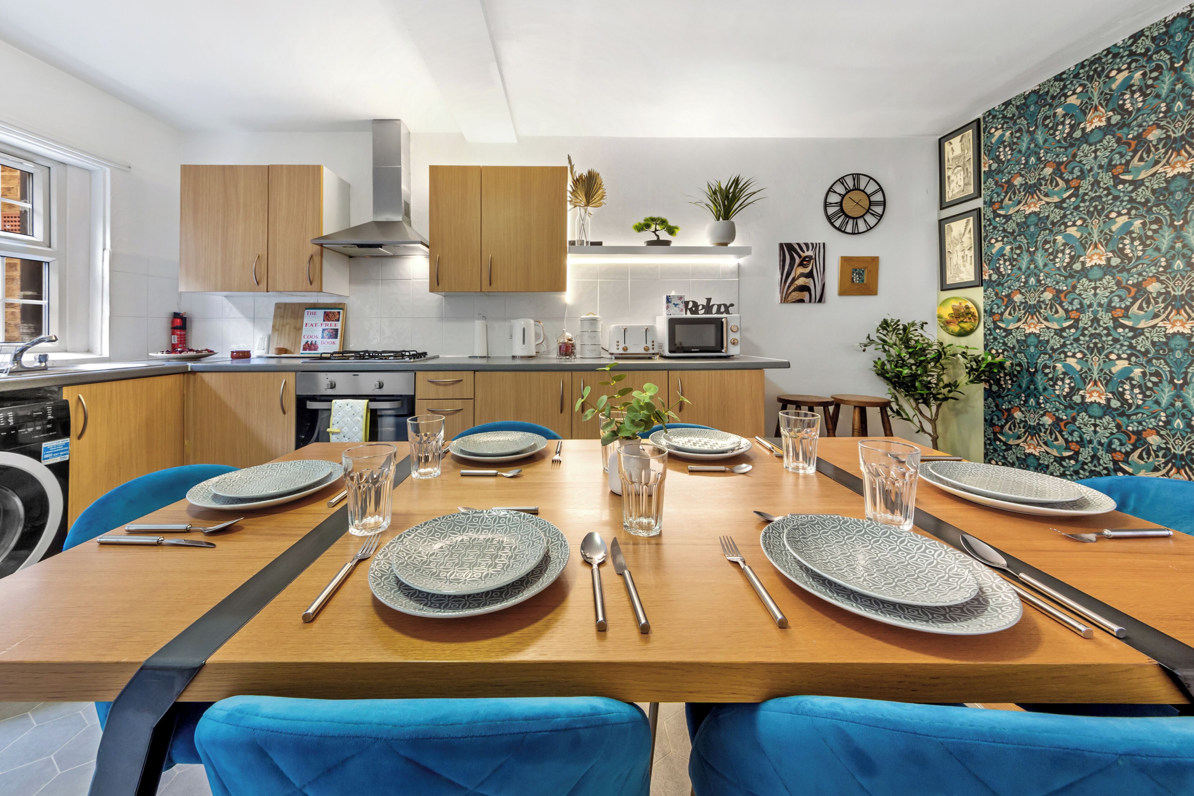 A modern kitchen with wooden cabinets, a dining table set for six, and a patterned wallpaper.