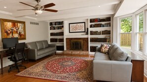 A living room with a gray sofa, a fireplace, bookshelves, a ceiling fan, and a large window. - Beautiful Lakeside Home 7 Minutes from Bellerive CC (Chesterfield)