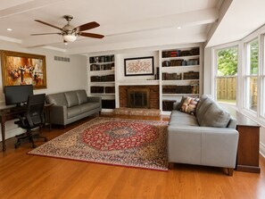 A living room with a gray sofa, a fireplace, bookshelves, a ceiling fan, and a large window. - Beautiful Lakeside Home 7 Minutes from Bellerive CC (Chesterfield)