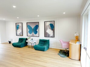 A modern living room with a wooden floor, two green sofas, a small round table, and a pink chair. There are three large butterfly paintings on the wall.