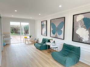 A spacious living area with a green sofa, a small round table, and a dining table with chairs. There are large framed butterfly artworks on the wall. - Shine Adam Court - Prestige Retreat in Pakenham (Pakenham)