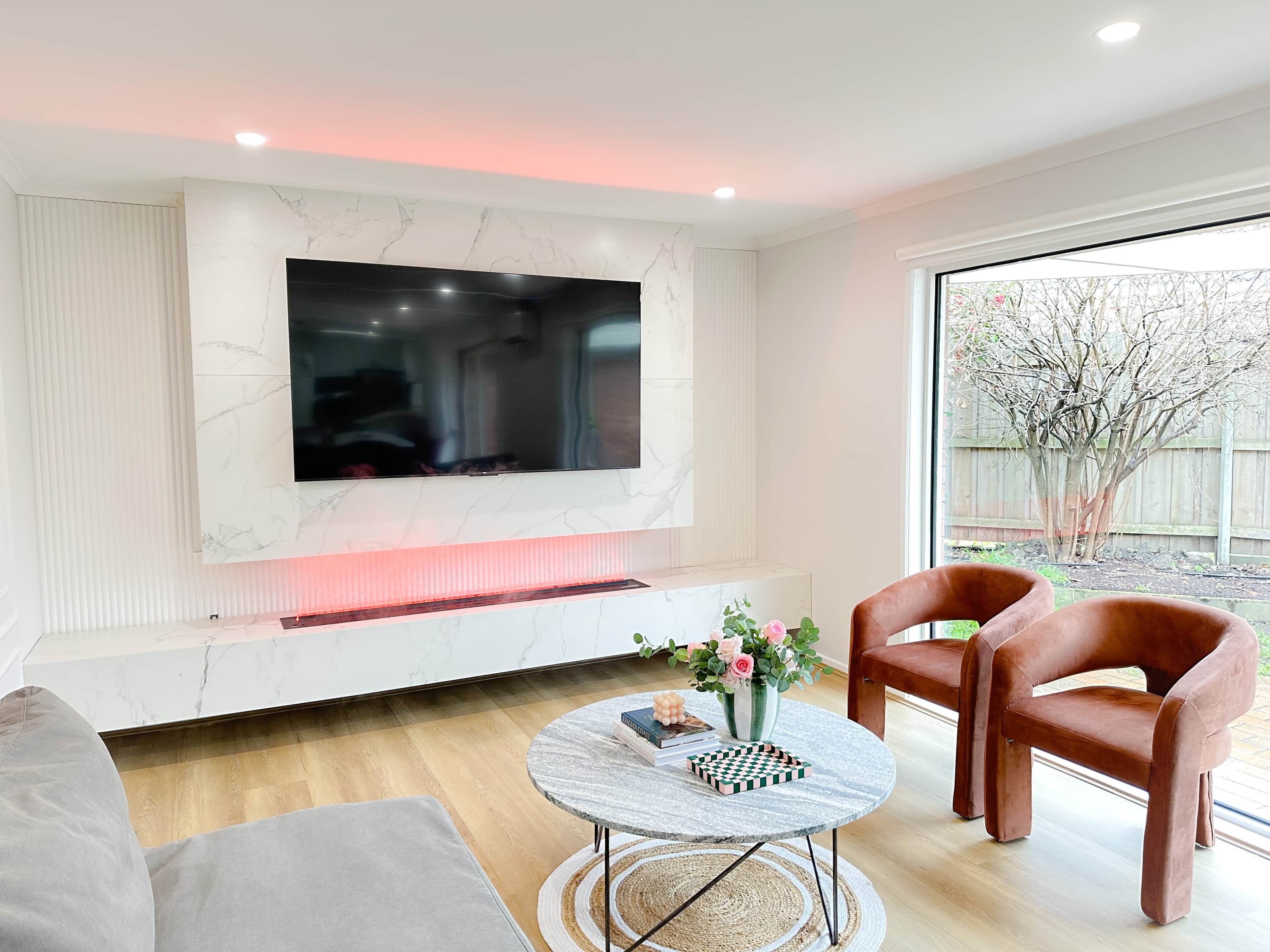A modern living room with a large flat-screen TV mounted on a marble wall, a round coffee table with books and a chessboard, two brown leather chairs, and a view of a tree through a large window.