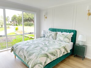 A bedroom with a large window, a bed with a green headboard, two bedside tables with lamps, and a view of a garden.