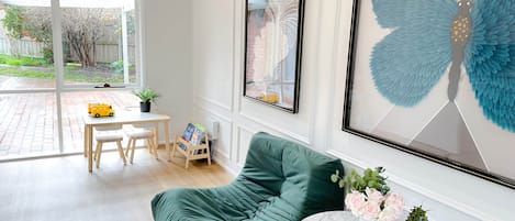 A room with a green sofa, a small round table with flowers, and a window with a view of a garden.