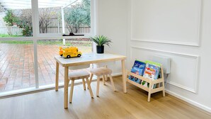 A room with a wooden table, two chairs, a potted plant, and a bookshelf with children's books. - Shine Adam Court - Prestige Retreat in Pakenham (Pakenham)