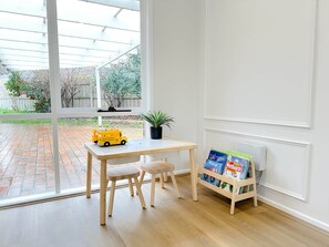 A room with a wooden table, two chairs, a potted plant, and a bookshelf with children's books. - Shine Adam Court - Prestige Retreat in Pakenham (Pakenham)