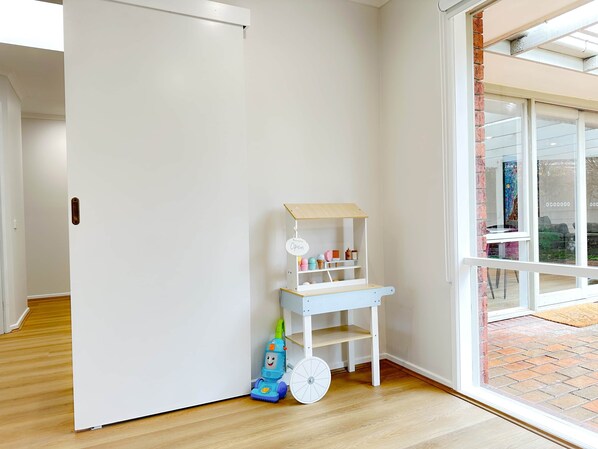A room with a white door, a small white shelf with toys, and a window with a view of a brick wall and a door.