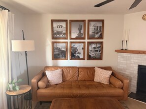 Ein Wohnzimmer mit braunem Ledersofa, einem Couchtisch und einem Kamin. An der Wand hängen sechs gerahmte Schwarz-Weiß-Fotos.