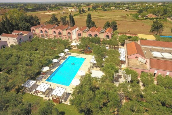 Luchtbeeld van een resort met een zwembad, omringd door ligbedden en parasols, een groot gebouw met rondboogvensters, en een landelijk landschap op de achtergrond.