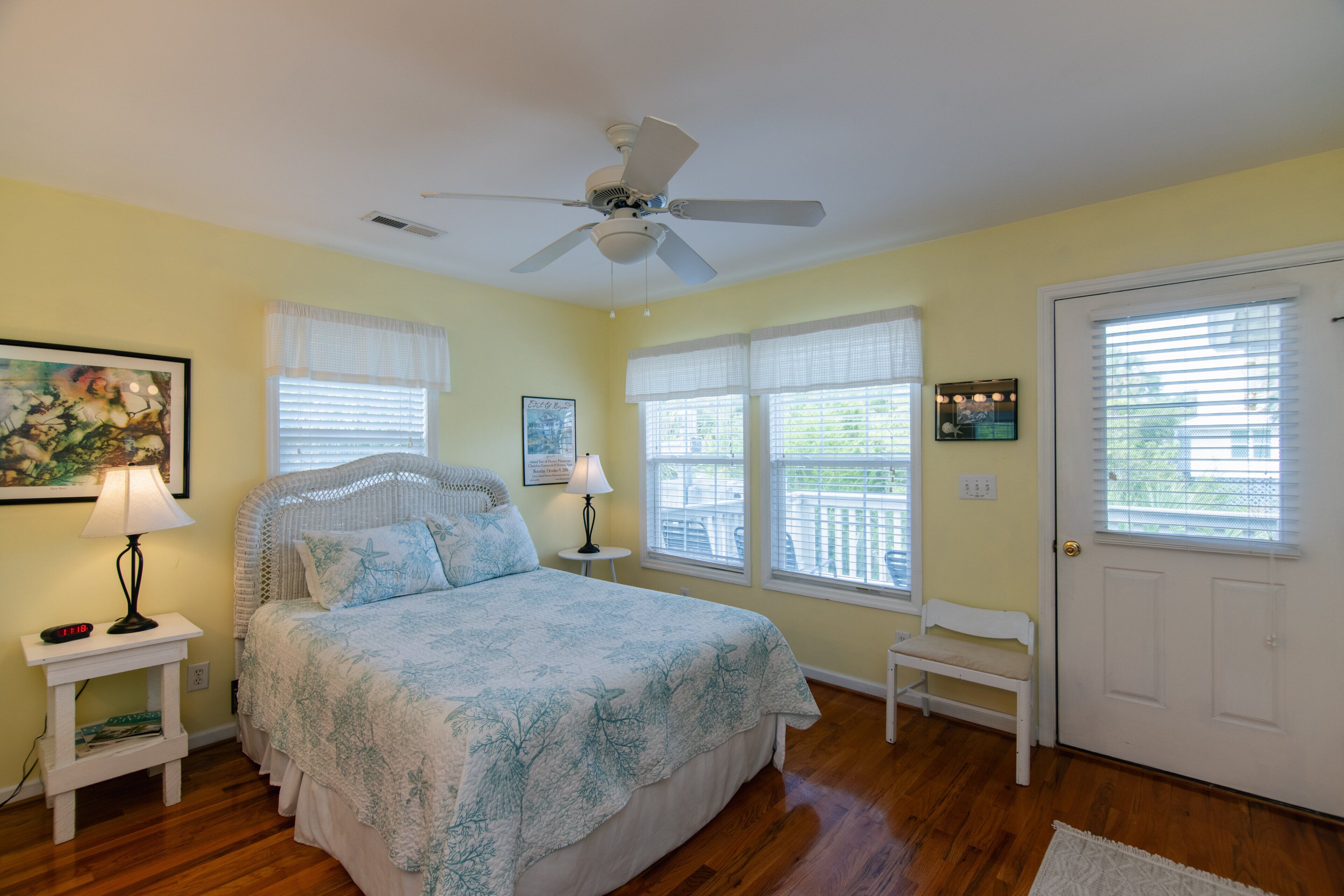Une chambre à coucher comprenant un lit, un ventilateur de plafond, une fenêtre avec des stores, une porte et une petite table avec une lampe.