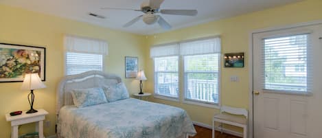 A bedroom with a bed, a ceiling fan, a window with blinds, a door, and a small table with a lamp.