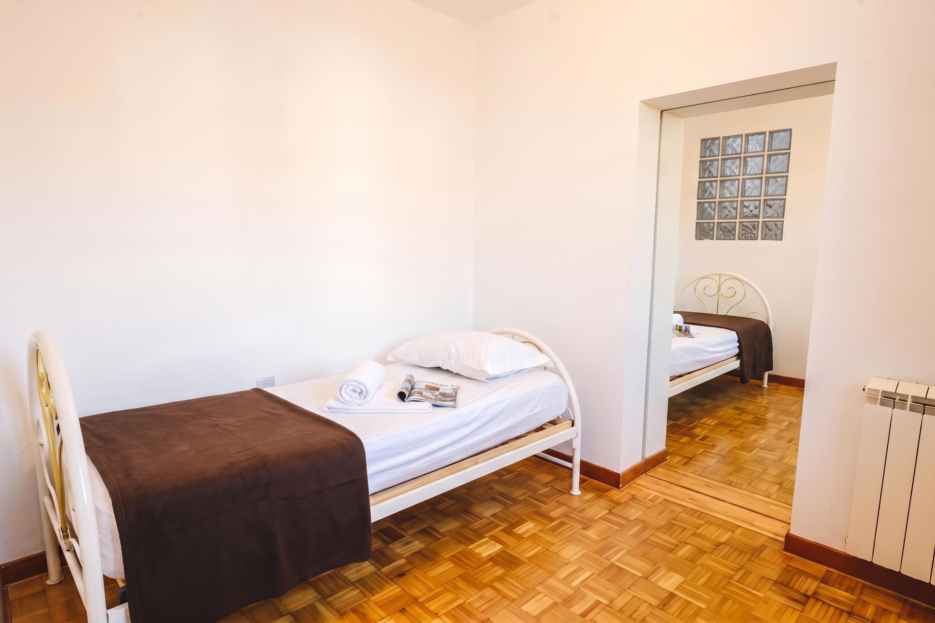 Uma cama de solteiro com um cobertor castanho, um travesseiro branco e uma revista sobre a cama. O chão é de madeira e há um radiador no lado direito.