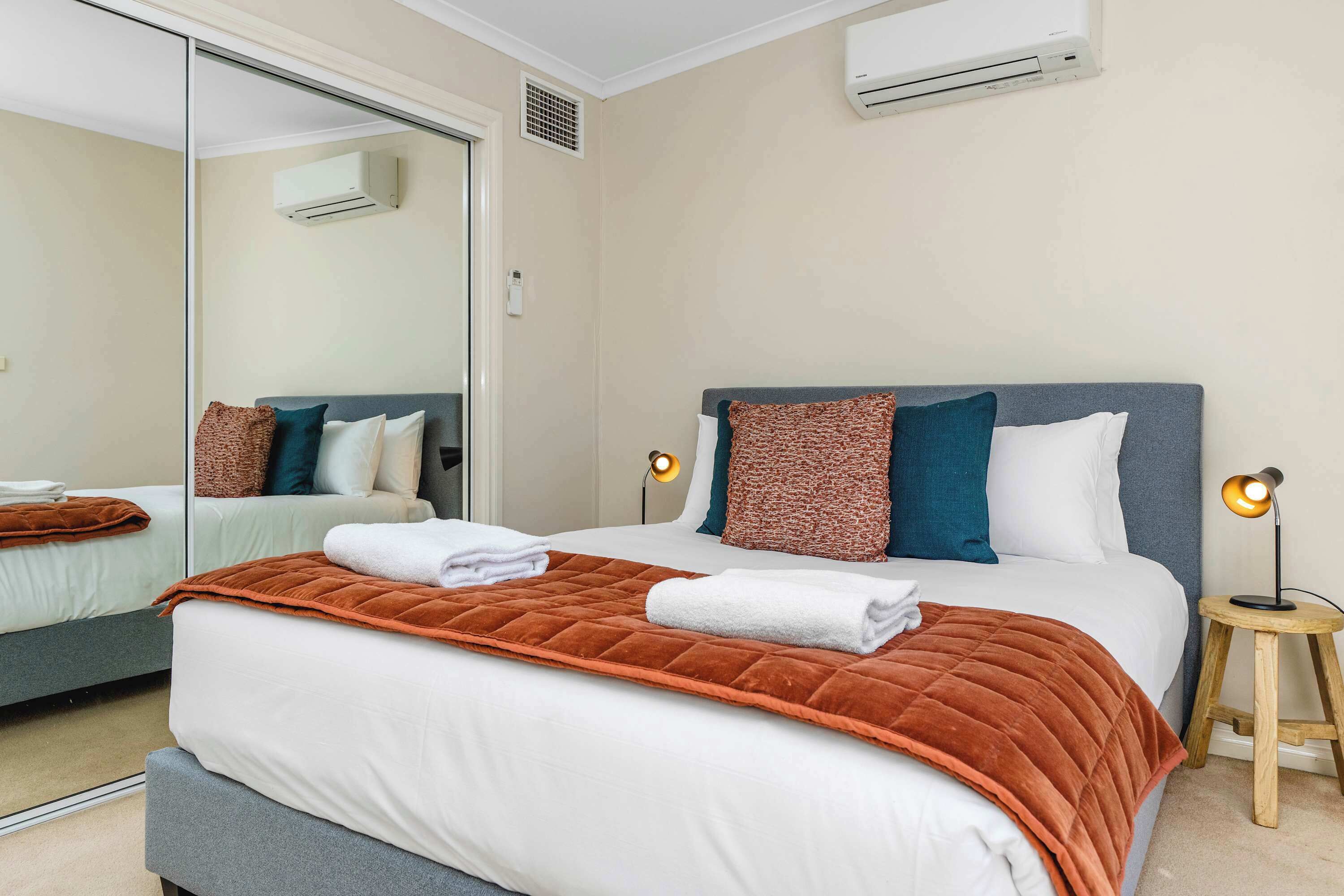 A hotel room with a bed, a bedside table with a lamp, a mirror, and an air conditioning unit on the wall.